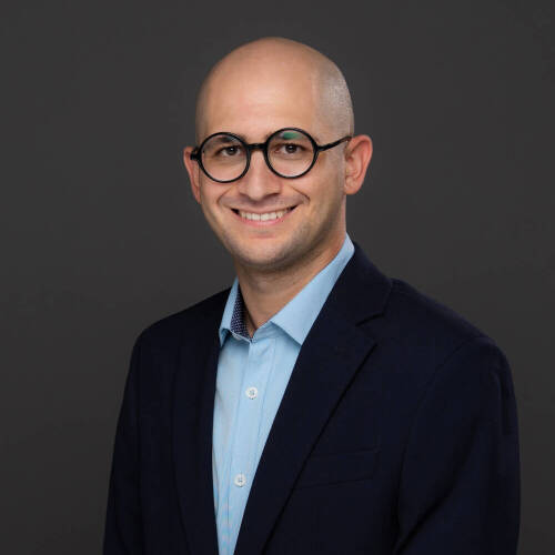 A person with a bald head is wearing a light blue collared shirt and a dark suit jacket, posed against a plain dark background.